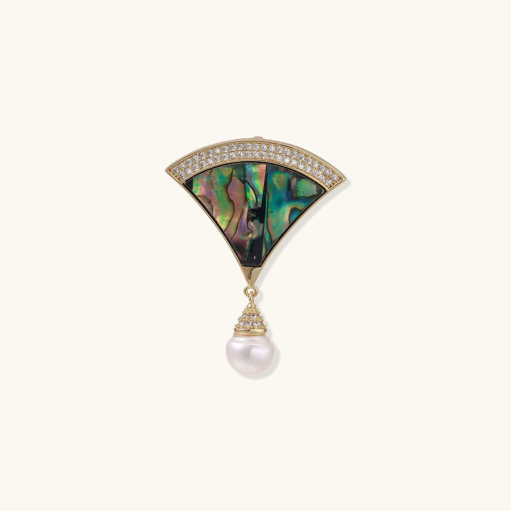 Gold fan-shaped brooch with iridescent abalone shell center, crystal border, and dangling white pearl on cream background