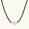 Beaded black agate necklace with iridescent baroque freshwater pearl centerpiece on cream background.